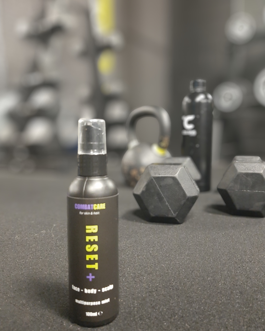 Black spray bottle labeled 'Combat Care Reset+' on a gym floor with weights in the background